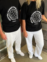 To personer iført sorte T-shirts med teksten "INVOLVING THE ENTIRE WORLD" og et cirkulært design. De står i en elevator med hvidt gulv og iført hvide bukser og sneakers.