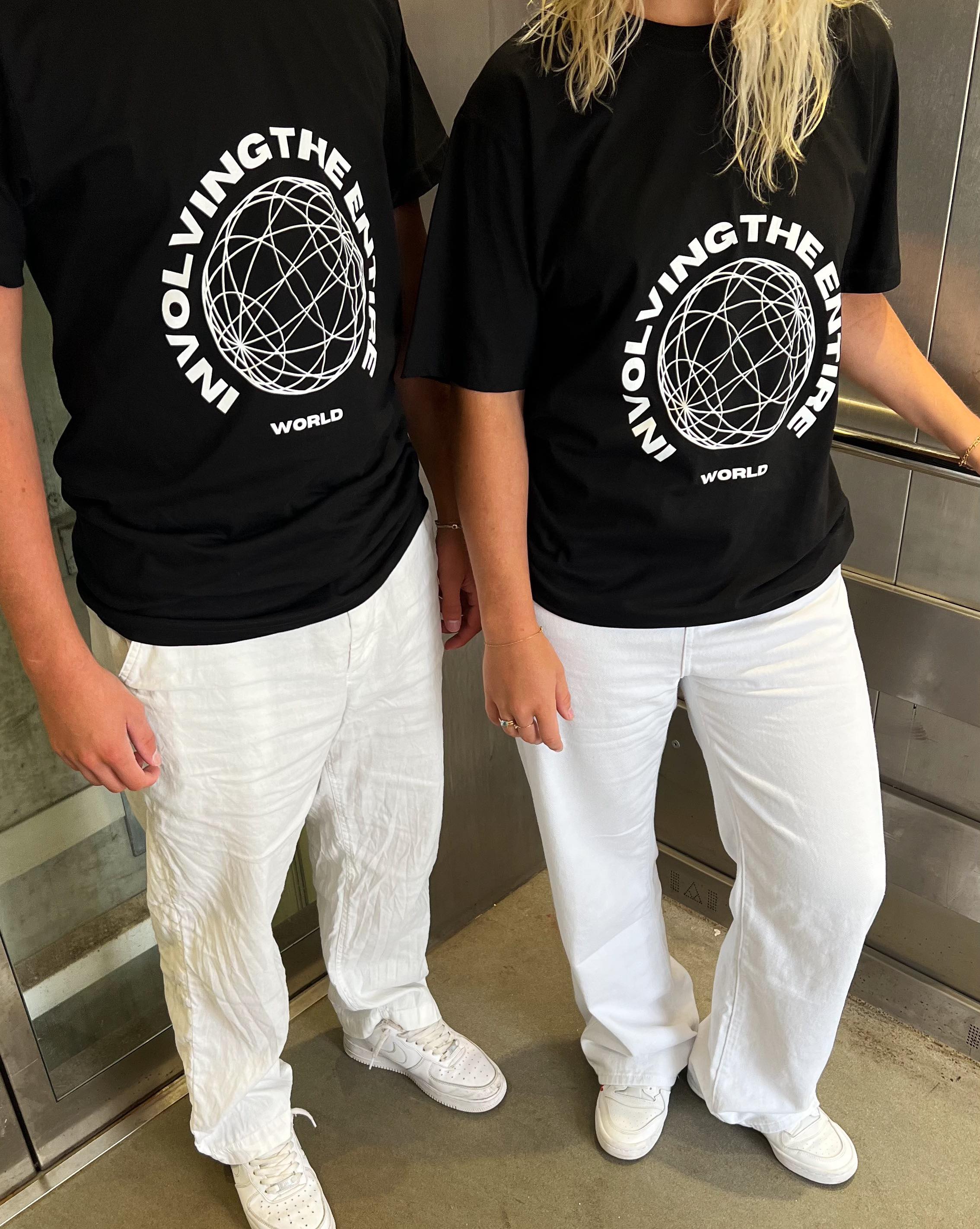 To personer iført sorte T-shirts med teksten "INVOLVING THE ENTIRE WORLD" og et cirkulært design. De står i en elevator med hvidt gulv og iført hvide bukser og sneakers.