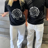 To personer iført sorte t-shirts med teksten "INVOLVING THE ENTIRE WORLD" står ved siden af hinanden, iført hvide bukser, i et rum med metallisk baggrund.