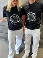 To personer iført sorte t-shirts med teksten "INVOLVING THE ENTIRE WORLD" står ved siden af hinanden, iført hvide bukser, i et rum med metallisk baggrund.