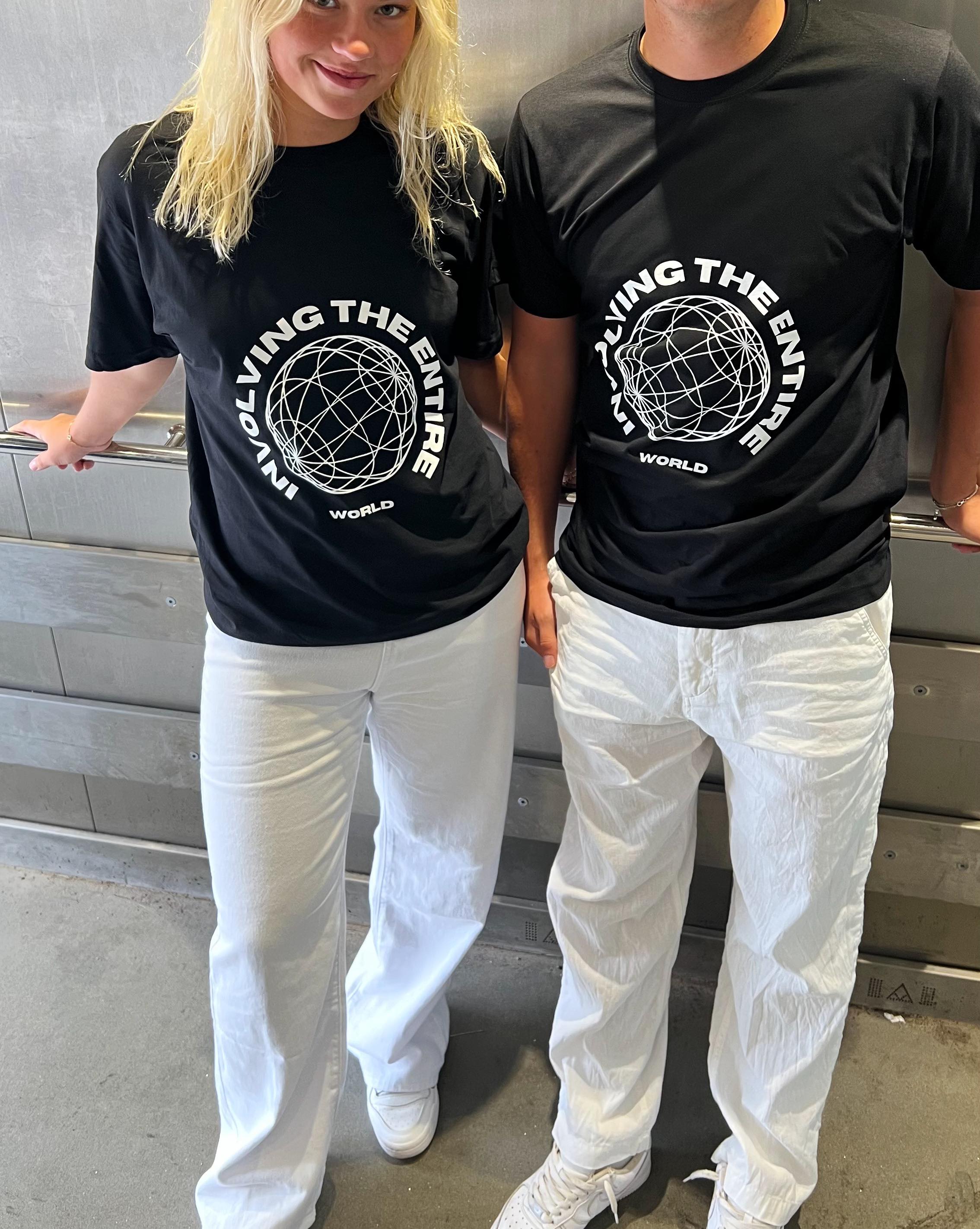 To personer iført sorte t-shirts med teksten "INVOLVING THE ENTIRE WORLD" står ved siden af hinanden, iført hvide bukser, i et rum med metallisk baggrund.