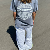 Person iført grå t-shirt med teksten “WORLDWIDE” over en globusgrafik, står på en asfaltvej under solen, iført hvide bukser.