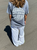 Person iført grå t-shirt med teksten “WORLDWIDE” over en globusgrafik, står på en asfaltvej under solen, iført hvide bukser.