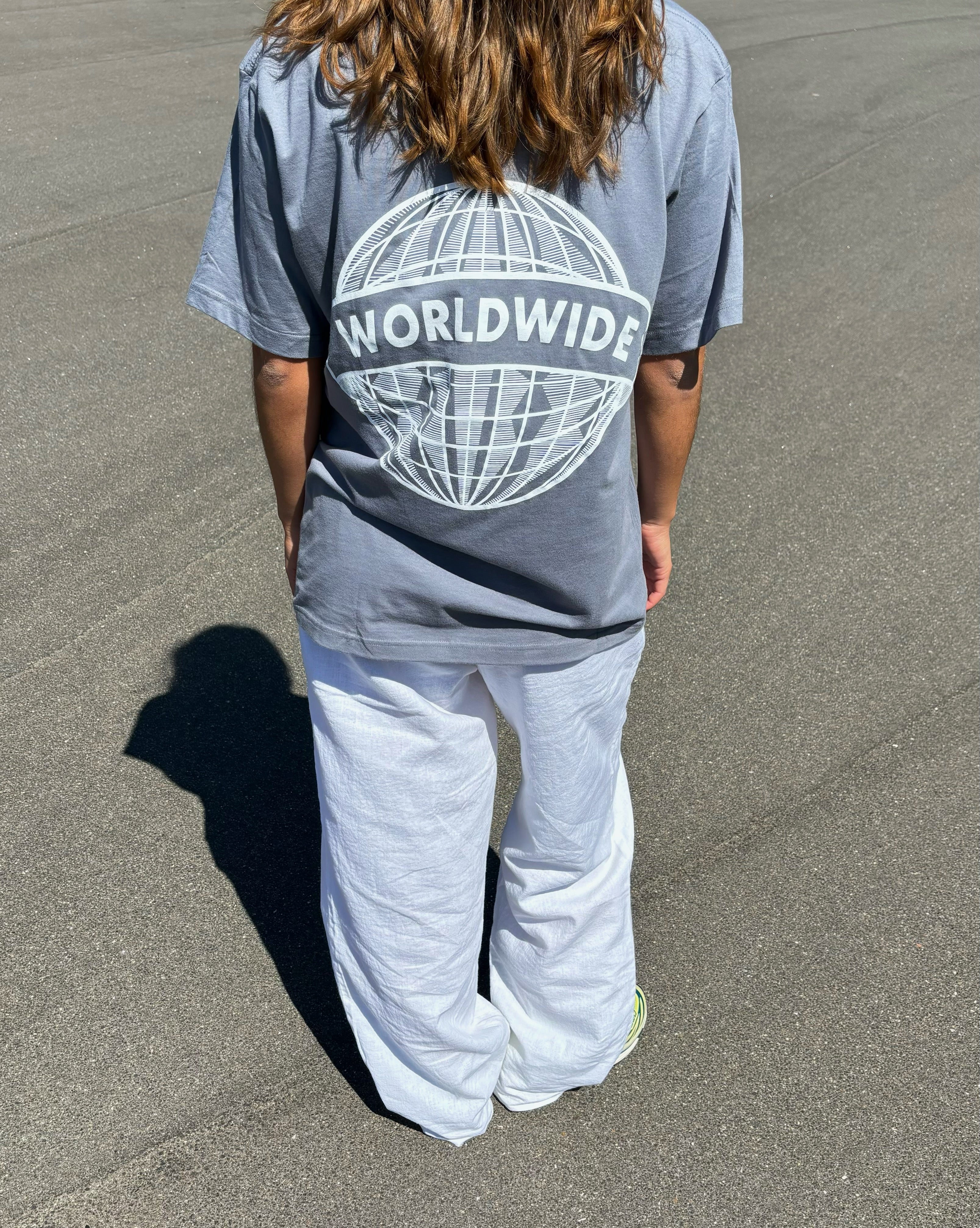 Person iført grå t-shirt med teksten “WORLDWIDE” over en globusgrafik, står på en asfaltvej under solen, iført hvide bukser.