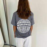 En person står med ryggen til iført en grå T-shirt med teksten "WORLDWIDE" og et globusdesign. Personen er i et minimalistisk rum med et spejl til venstre.