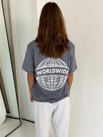 En person står med ryggen til iført en grå T-shirt med teksten "WORLDWIDE" og et globusdesign. Personen er i et minimalistisk rum med et spejl til venstre.