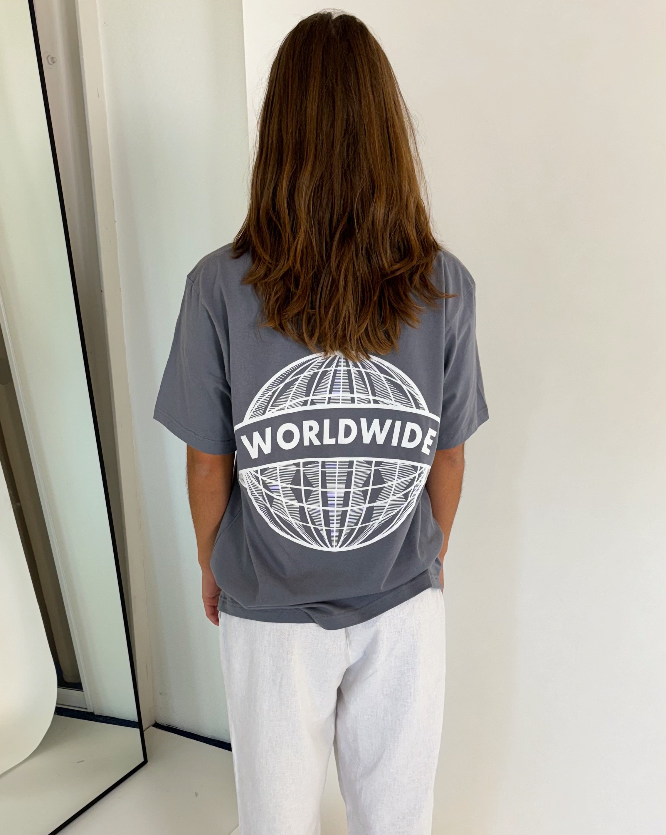 En person står med ryggen til iført en grå T-shirt med teksten "WORLDWIDE" og et globusdesign. Personen er i et minimalistisk rum med et spejl til venstre.