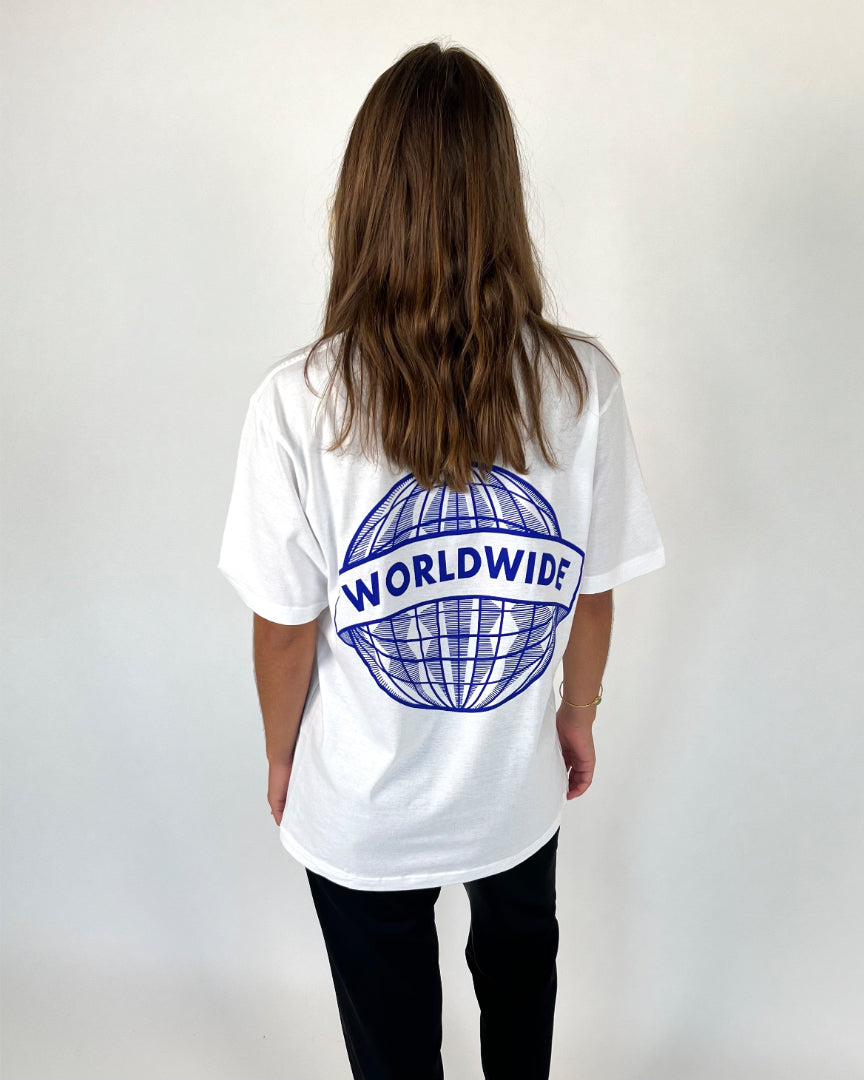 En person iført en hvid t-shirt med et blåt globusmotiv og teksten "WORLDWIDE" står med ryggen til kameraet i et neutralt, lyst rum.