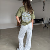 En person iført en olivengrøn T-shirt med teksten "WORLDWIDE" og en globusdesign står i et minimalistisk rum med beton- og hvidmalede overflader, omgivet af møbler.