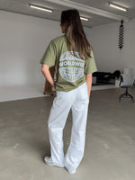 En person iført en olivengrøn T-shirt med teksten "WORLDWIDE" og en globusdesign står i et minimalistisk rum med beton- og hvidmalede overflader, omgivet af møbler.