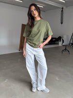 En person i en grøn t-shirt og hvide bukser står afslappet på et råt betongulv i et lyst studie, med fotoudstyr og en stol i baggrunden.
