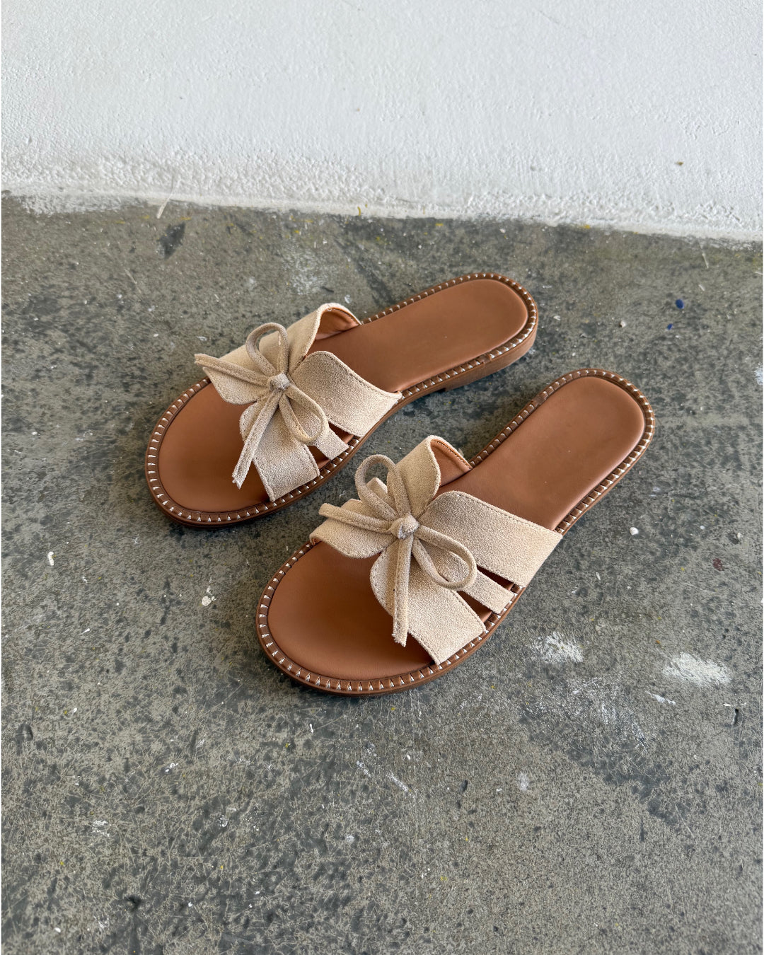 Beige sandaler med sløjfer ligger på et slidt betongulv, omgivet af en hvid væg.