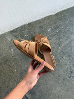 En hånd holder et par lysebrune sandaler med sløjfer, mens den står på en grå betonoverflade.