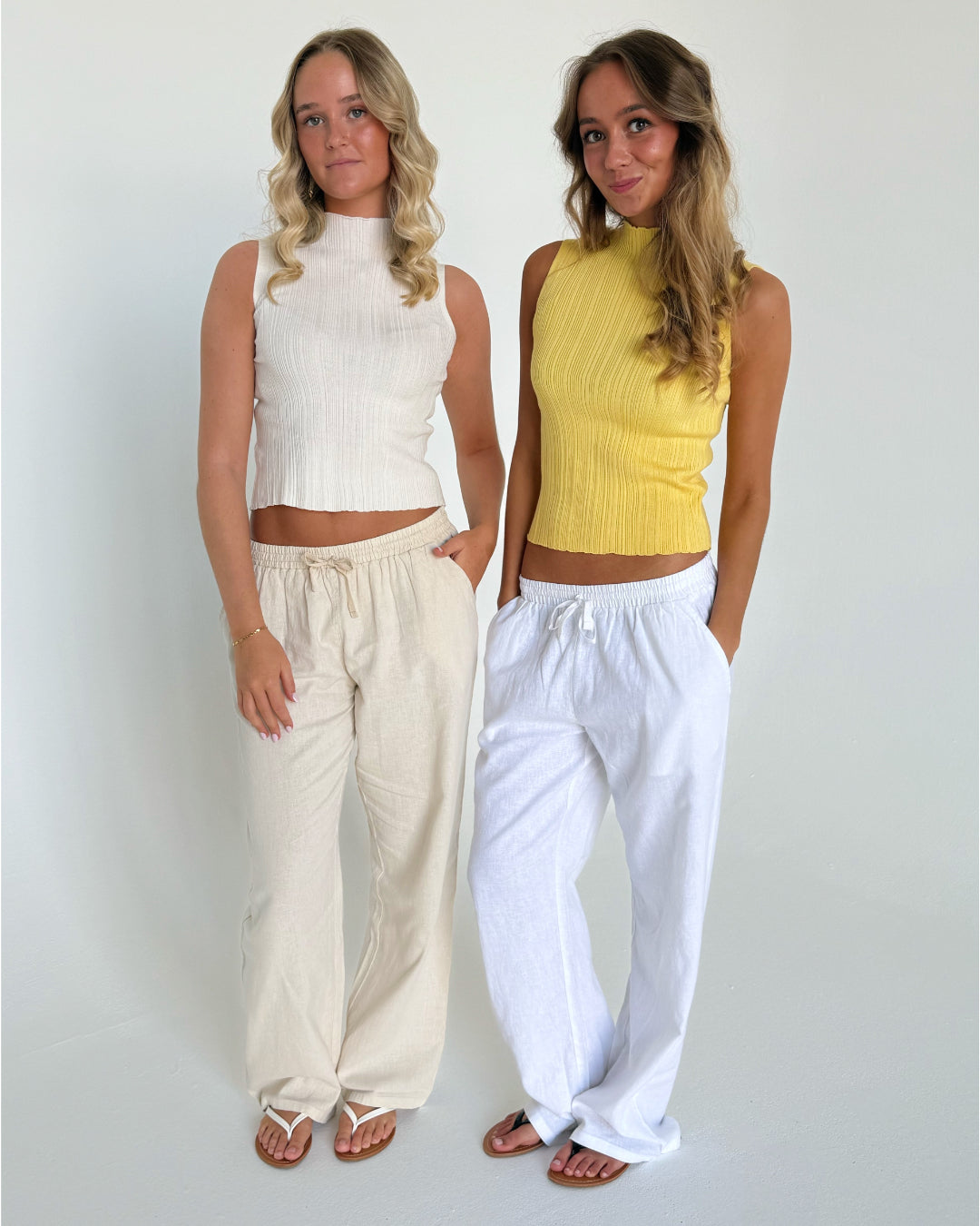 To personer står sammen, iført lyse sommeroutfits med løse bukser og ærmeløse toppe; den ene i gule og hvide nuancer, den anden i nude nuancer. Baggrunden er en enkel, hvid væg.