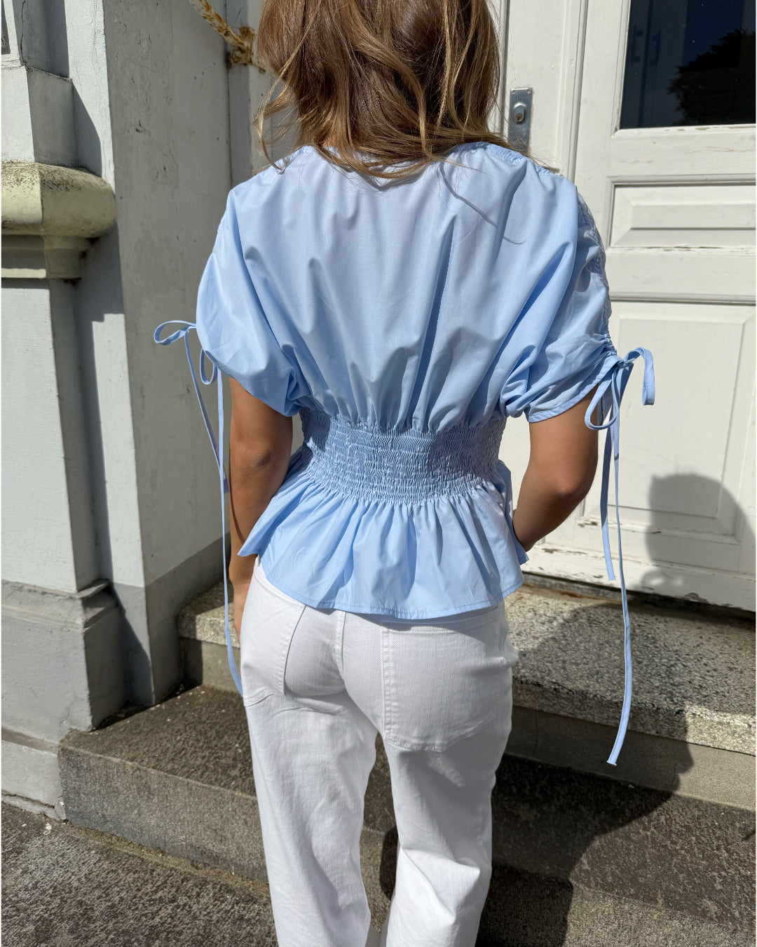 En person iført en lyseblå bluse og hvide bukser står på en trappe foran en hvid dør, omgivet af lyse murstensvægge.