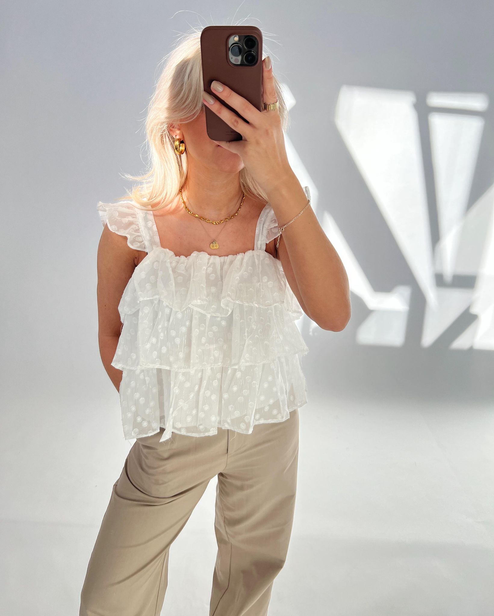 Person holder en mobiltelefon foran ansigtet, iført en hvid, ruffled bluse og beige bukser. Omgivet af skygger i et lyst rum.