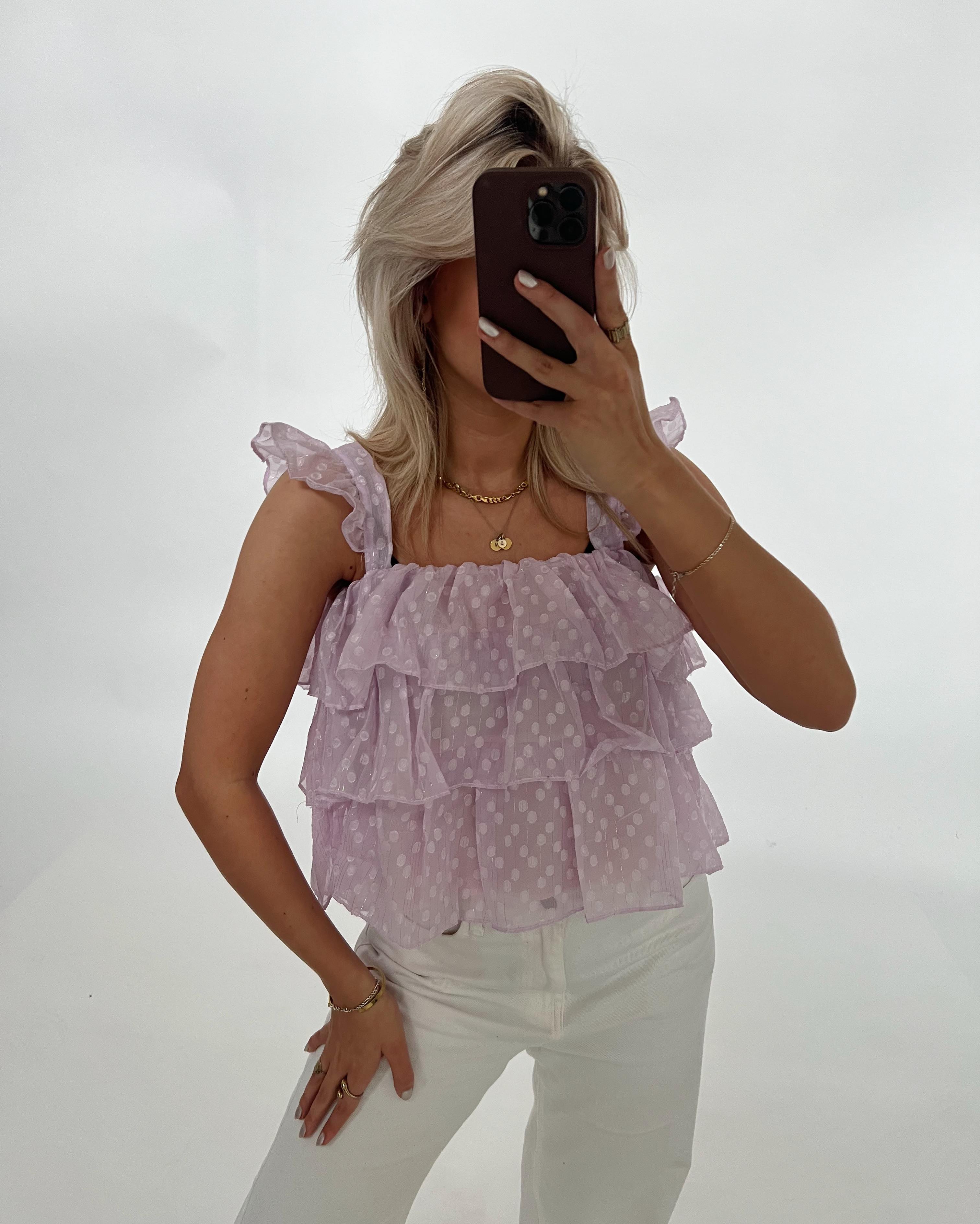 Kvinde i lyslilla, prikket, flæsebluse tager selfie; holdning med hånden på hoften, bærer hvide bukser og smykker; neutral baggrund.