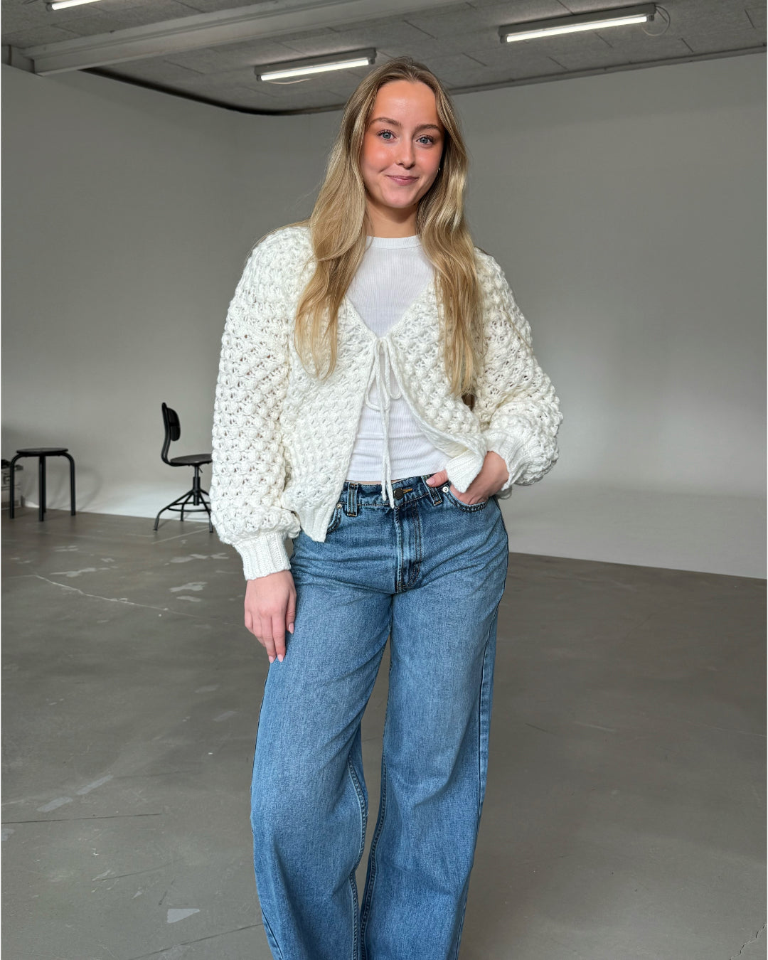 En person iført en hvid strikket cardigan og blå jeans står smilende i et minimalistisk rum med grå gulve og hvide vægge. I baggrunden er der sorte stole. Ingen tekst.