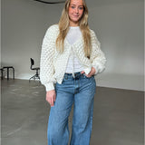En person iført en hvid strikket cardigan og blå jeans står smilende i et minimalistisk rum med grå gulve og hvide vægge. I baggrunden er der sorte stole. Ingen tekst.