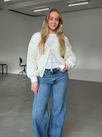 En person iført en hvid strikket cardigan og blå jeans står smilende i et minimalistisk rum med grå gulve og hvide vægge. I baggrunden er der sorte stole. Ingen tekst.