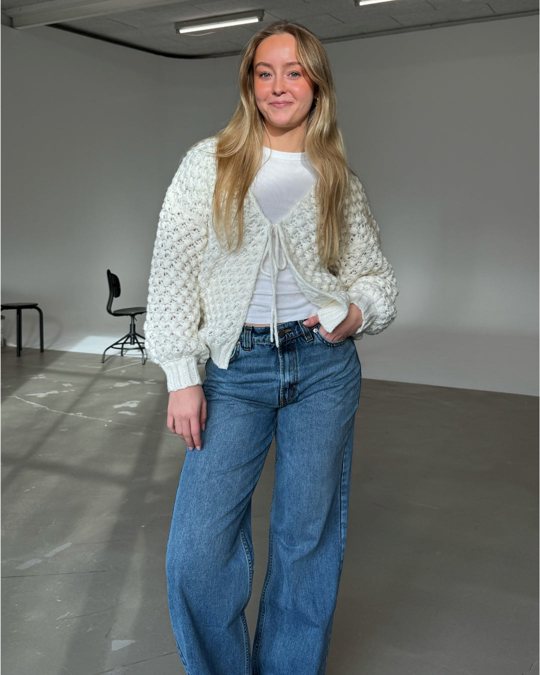 Kvinde står med et smil iført en strikket cardigan og jeans i et enkelt rum med grå gulv og sorte stole i baggrunden.