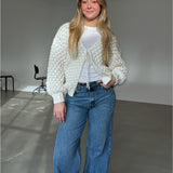 Kvinde står med et smil iført en strikket cardigan og jeans i et enkelt rum med grå gulv og sorte stole i baggrunden.