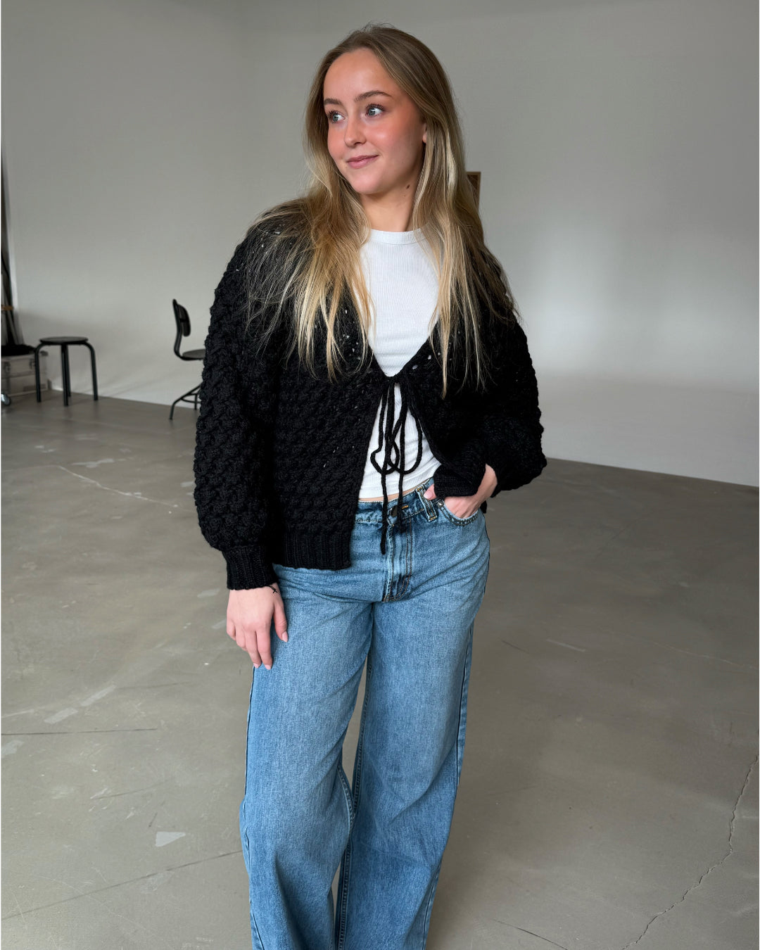 En kvinde iført en sort striktrøje og blå jeans står afslappet og kigger til siden i et minimalistisk rum med gråt gulv og få møbler.