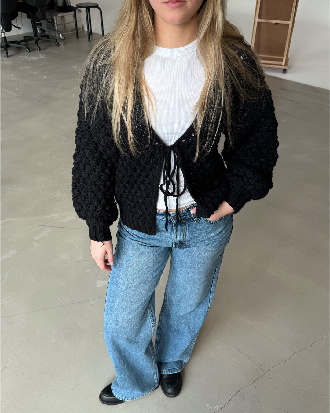 En person står iført en sort strikket cardigan og blå jeans, hviler en hånd i lommen, i et minimalistisk kontorrum med lysegråt gulv og stole i baggrunden.
