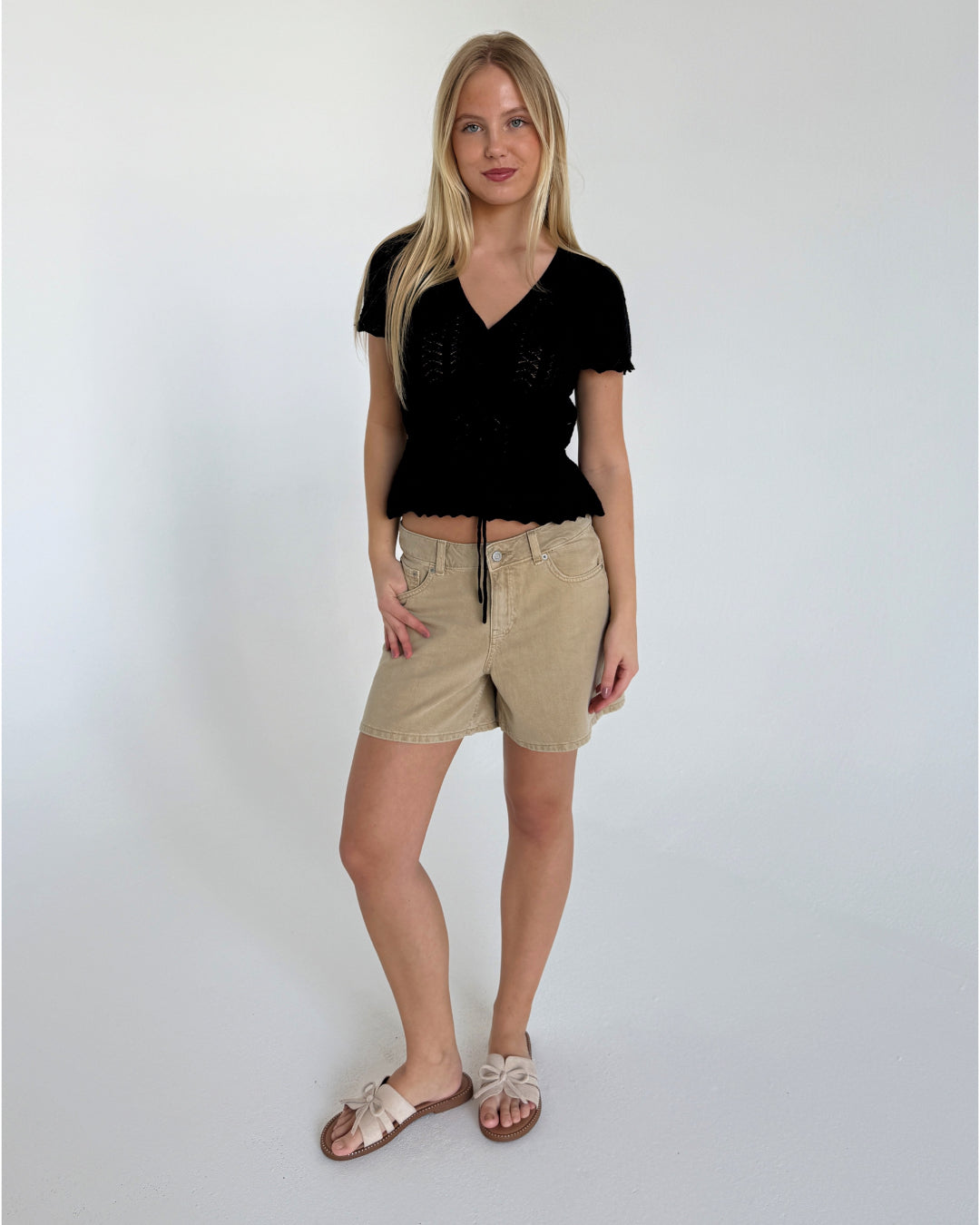 En kvinde iført sort bluse og beige shorts står i et minimalistisk, hvidt rum. Hun har lange, blondehår og sandaler med sløjfe, og poserer afslappet.