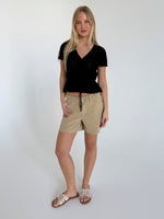 En kvinde iført sort bluse og beige shorts står i et minimalistisk, hvidt rum. Hun har lange, blondehår og sandaler med sløjfe, og poserer afslappet.