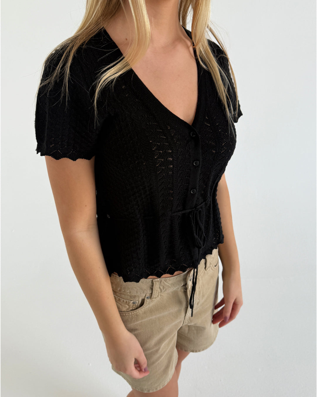 En person bærer en sort, strikket bluse med korte ærmer og beige shorts. Personen står indendørs mod en ren, hvid væg.
