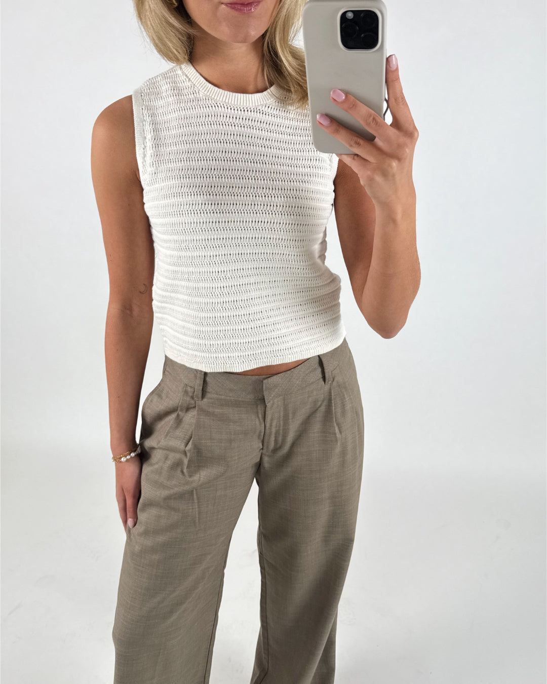 En person iført en hvid strikket top og beige bukser tager en selfie med en smartphone. Personen står i et neutralt, hvidt rum. Ingen tekst til stede.