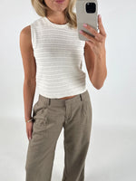 En person iført en hvid strikket top og beige bukser tager en selfie med en smartphone. Personen står i et neutralt, hvidt rum. Ingen tekst til stede.