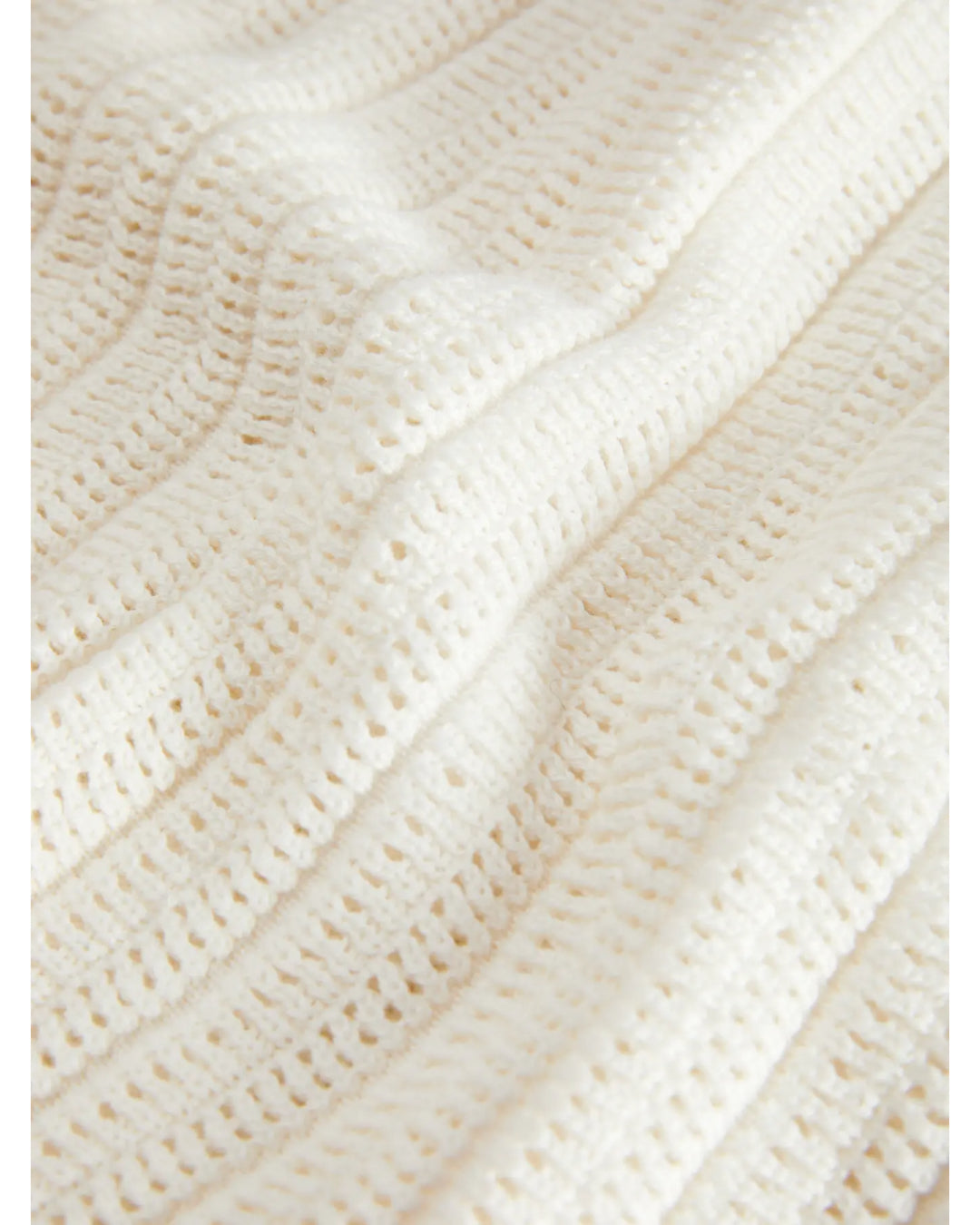 Et stykke flødefarvet strikstof ligger i bølgete folder. Stoffet består af et åbent hæklet mønster, som giver det en luftig og tekstureret fremtoning.